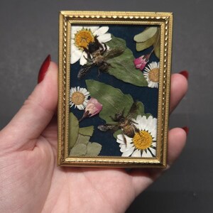 May include: A small rectangular gold-toned frame containing a dark blue velvet backing. The display features two preserved bees positioned among pressed white daisy flowers, green leaves, and small pink flower petals arranged on the surface.