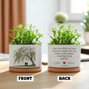May include: Two white ceramic planters with green plants. One planter features a willow tree illustration and two figures. The other planter has text that reads "Like the willow...standing strong together." Includes names and a date.
