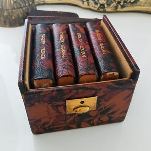 Puede incluir: Una pequeña caja de cuero vintage con cuatro libros en miniatura. Cada libro está etiquetado con "English" y un idioma diferente: francés, alemán, español e italiano. La caja tiene un cierre de latón y un exterior estampado. Una figura decorativa y una pintura enmarcada están en el fondo.
