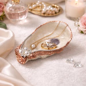 May include: An oyster shell trinket dish with a metallic copper-toned rim and a cluster of white faux pearls and clear crystals on one side. The interior of the shell is iridescent and holds a gold-toned necklace with a pearl pendant and a gold ring.