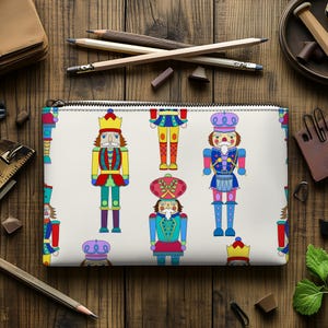 May include: A white pencil case featuring a repeating pattern of colorful nutcracker figures. The nutcrackers are depicted in various poses, wearing crowns and uniforms in shades of red, yellow, blue, and green. The case has a zipper closure.