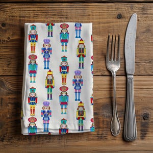 May include: White cloth napkin with a repeating pattern of colorful nutcracker figures. The nutcrackers are in various colors, including red, blue, yellow, and purple. A fork and knife are placed to the right of the napkin on a wooden surface.