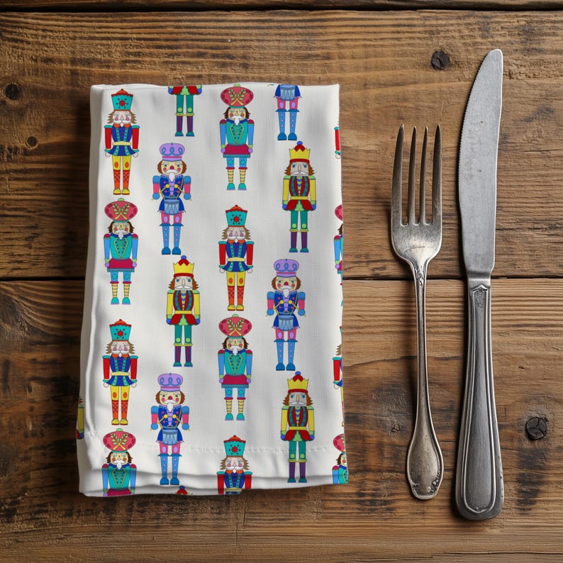 May include: White cloth napkin with a repeating pattern of colorful nutcracker figures. The nutcrackers are in various colors, including red, blue, yellow, and purple. A fork and knife are placed to the right of the napkin on a wooden surface.