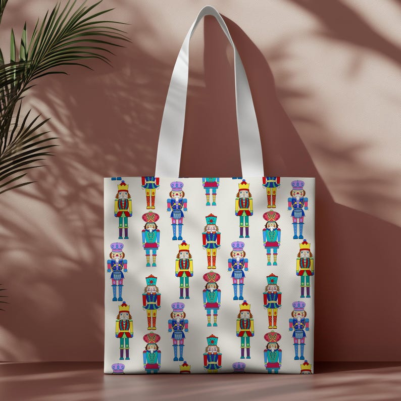 May include: A cream-colored tote bag featuring a repeating pattern of colorful nutcracker figures. The nutcrackers are depicted in various uniforms with crowns, hats, and jackets in shades of blue, red, yellow, and green. The bag has white handles.
