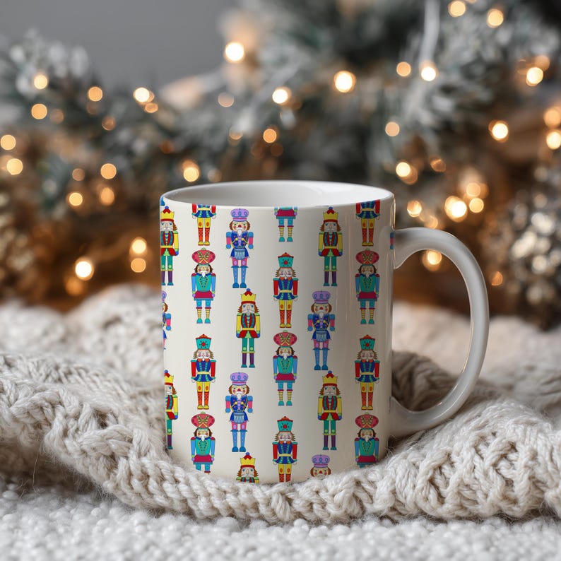 May include: White ceramic mug featuring a repeating pattern of colorful nutcracker figures. The nutcrackers are depicted in various uniforms with details in red, blue, yellow, and green. The mug has a white handle and is set against a blurred background.