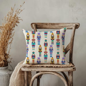 May include: A decorative square pillow featuring a repeating pattern of colorful nutcracker soldiers. The nutcrackers are in various colors, including red, blue, green, and yellow, on a cream-colored background. The pillow is placed on a wooden chair.