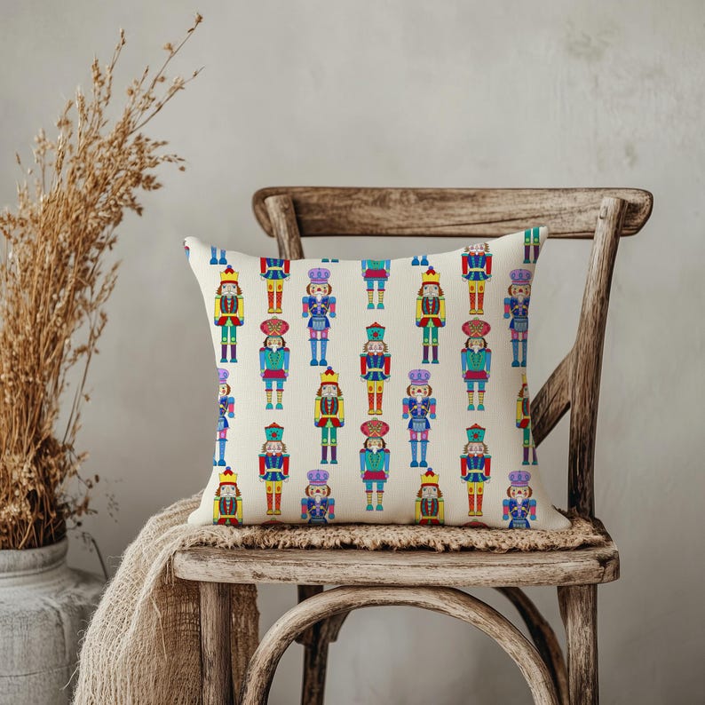May include: A decorative square pillow featuring a repeating pattern of colorful nutcracker soldiers. The nutcrackers are in various colors, including red, blue, green, and yellow, on a cream-colored background. The pillow is placed on a wooden chair.