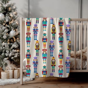 May include: A cream-colored blanket featuring a repeating pattern of colorful nutcracker figures. The nutcrackers are depicted in various poses and outfits, with details in red, blue, yellow, and green. The blanket is draped over a crib.
