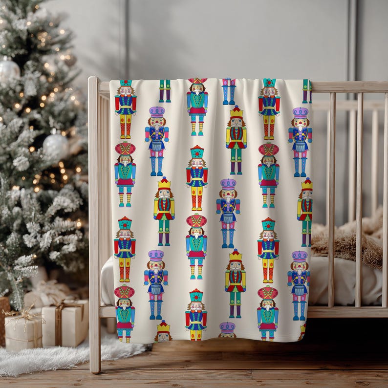 May include: A cream-colored blanket featuring a repeating pattern of colorful nutcracker figures. The nutcrackers are depicted in various poses and outfits, with details in red, blue, yellow, and green. The blanket is draped over a crib.