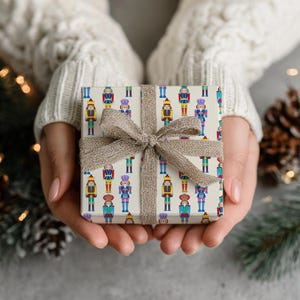 May include: A wrapped gift box featuring a repeating pattern of colorful nutcracker soldiers on a cream background. The box is tied with a burlap ribbon. The gift is held in two hands, suggesting a holiday or special occasion.