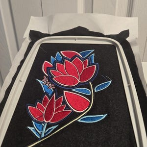 May include: Embroidered floral design featuring two red lotus flowers with blue accents on a black felt background. The design is held in a white embroidery hoop, with a layer of white stabilizer on top.