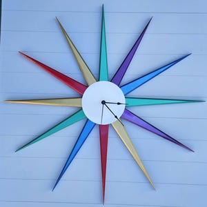 May include: A starburst wall clock with a white circular centre and colourful, pointed rays in red, gold, teal, blue, purple, and green. The clock hands are black.