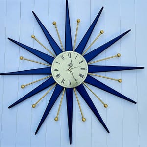 May include: A navy blue starburst wall clock with gold detailing. The clock face is cream-coloured with black Roman numerals and hands. The starburst design features long, pointed navy blue rays with gold spheres.