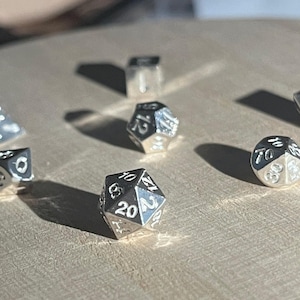 May include: A selection of silver polyhedral dice, including a D4, D6, D8, D10, D12, D20, and a percentile die. The dice are arranged on a wooden surface, each showing its numerical face. These dice are for tabletop games.
