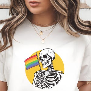 Könnte beinhalten: Weißes T-Shirt mit einem schwarz-weißen Skelett, das eine Regenbogenflagge vor einem gelben Kreis hält. Der Text "Digital Download 300 DPI PNG file!" befindet sich am unteren Rand des Bildes.