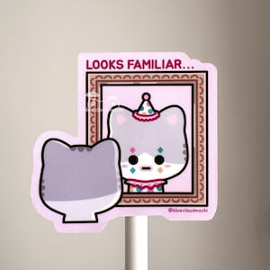 Peut inclure: Un autocollant découpé représentant une tête de chat violet clair au premier plan et le reflet dans un miroir encadré d'un chat portant un chapeau de clown coloré, une collerette et du maquillage en losange. Le texte LOOKS FAMILIAR est en rose.