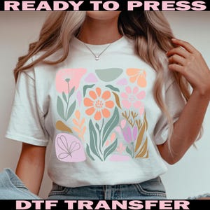 May include: White t-shirt featuring a colorful floral design with pink, orange, and green elements. The words "READY TO PRESS" and "DTF TRANSFER" are printed in pink and black, respectively, at the top and bottom of the image.
