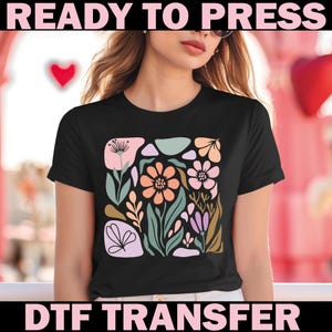 May include: Black t-shirt featuring a colorful floral design with peach, pink, green, and purple elements. The words "READY TO PRESS" and "DTF TRANSFER" are printed in pink above and below the design. The shirt has short sleeves.