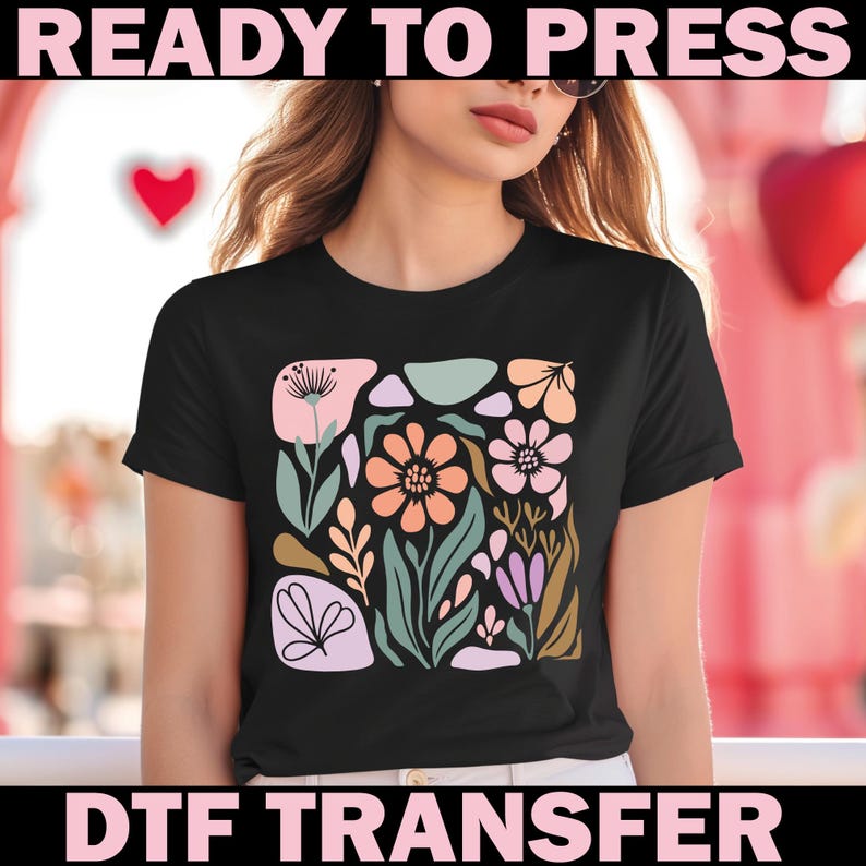 May include: Black t-shirt featuring a colorful floral design with peach, pink, green, and purple elements. The words "READY TO PRESS" and "DTF TRANSFER" are printed in pink above and below the design. The shirt has short sleeves.
