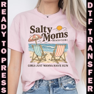 May include: A light pink t-shirt featuring a beach scene with two striped beach chairs, an umbrella, and the text "Salty Moms Beach Club." The phrase "Girls Just Wanna Have Sun" is also printed on the shirt.