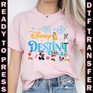 K&ouml;nnte beinhalten: Hellrosa T-Shirt mit dem Schriftzug "Disney Destiny" in Blau und Gelb, mit Cartoonfiguren wie Mickey und Minnie Mouse. Das Design enth&auml;lt auch Feuerwerk und Sterne.
