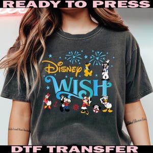 K&ouml;nnte beinhalten: Dunkelgraues T-Shirt mit den Worten "Disney Wish" in Gelb und Blau sowie Disney-Figuren. Das Shirt tr&auml;gt auch die Worte "READY TO PRESS" und "DTF TRANSFER" in Pink.