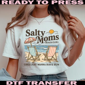 May include: White t-shirt with a beach-themed graphic. The design features the text "Salty Moms Beach Club" with an illustration of beach chairs, an umbrella, and ocean waves. The phrase "GIRLS JUST WANNA HAVE SUN" is also included. The words "READY TO PRESS" and "DTF TRANSFER" are also included.