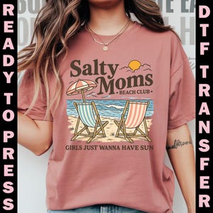 May include: A dusty rose t-shirt featuring a beach scene with two beach chairs, an umbrella, and the text "Salty Moms Beach Club." The phrase "Girls Just Wanna Have Sun" is also printed on the shirt. The words "Ready to Press" and "DTF Transfer" are printed on the sides.