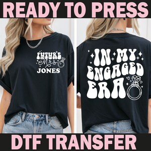 May include: Black t-shirt with white text and graphics. The front reads "Future Mrs. Jones" with a ring graphic. The back says "In My Engaged Era" with a ring graphic. The words "Ready to Press" and "DTF Transfer" are at the top and bottom.