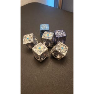 May include: Six faceted dice in shades of blue and silver are arranged on a dark surface. Each die has a central design with the text "BLUE DOT". The dice are cube-shaped and appear to be made of a clear material.