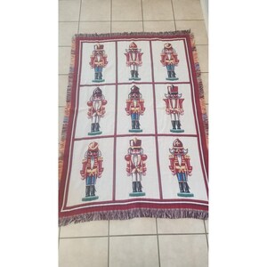 May include: A rectangular woven throw blanket featuring a grid pattern with nine distinct nutcracker figures. Each nutcracker is depicted in red, blue, and gold uniforms against a white background, framed by dark red borders with fringed edges.