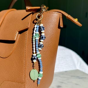 May include: A brown leather handbag with a gold-colored chain and a beaded charm. The charm features blue, white, and green beads, including a large, light green, oval-shaped stone. The bag has a top handle and a flap closure.