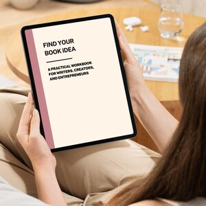 Pode incluir: Um tablet exibe o texto "FIND YOUR BOOK IDEA" com o subtítulo "A PRACTICAL WORKBOOK FOR WRITERS, CREATORS, AND ENTREPRENEURS". O tablet tem uma moldura preta e uma faixa vertical rosa. O fundo é bege suave.