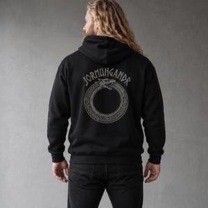 Puede incluir: Una sudadera negra con capucha que presenta un gráfico gris en la parte posterior. El diseño muestra una serpiente circular mordiéndose la cola, un Ouroboros, con el texto JORMUNGANDR arqueado sobre la serpiente en una fuente negrita.