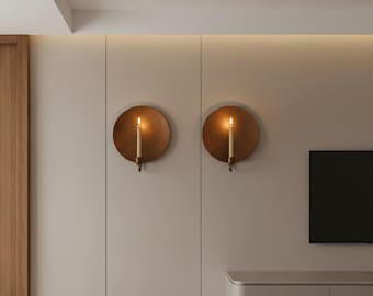 May include: Two round, antique brass wall sconces with lit candles. Each sconce has a circular backplate and a curved arm holding a single candle. The sconces are mounted on a light beige wall.