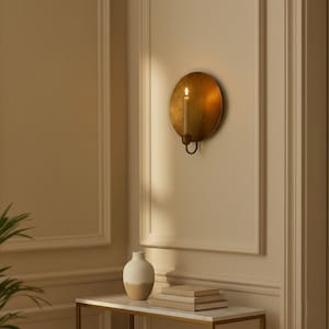 May include: A wall-mounted candle holder with a lit candle. The holder is a round, antique gold color. The candle is a light beige color. A small table with a vase and books is below the candle holder.