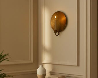 May include: A wall-mounted candle holder with a lit candle. The holder is a round, antique gold color. The candle is a light beige color. A small table with a vase and books is below the candle holder.