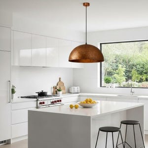 Puede incluir: Una lámpara colgante de color cobre cuelga sobre una isla de cocina blanca. La lámpara tiene forma de cúpula con una superficie texturizada. La cocina cuenta con gabinetes blancos, una estufa de acero inoxidable y una ventana grande.