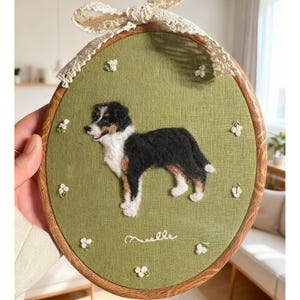 以下が含まれることがあります： 黒、白、茶色の毛並みの犬が描かれた刺繍アート。緑色の背景に、木製のフレームに収められ、小さな白い花のアクセントとレースのリボンで飾られています。