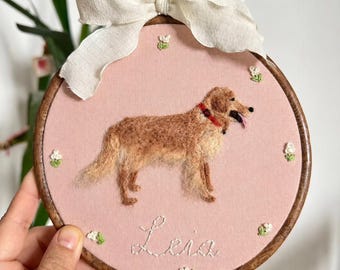 May include: A circular wooden embroidery hoop containing pink fabric with a needle-felted golden dog wearing a red collar. The name "Leia" is embroidered in white script below the dog, surrounded by small white floral accents and a white ribbon.