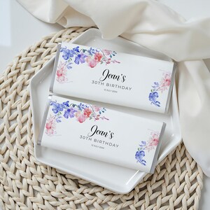 May include: Two white chocolate bar wrappers with floral watercolor designs in pink, purple, and blue. The text on the wrappers reads "Jean's 30th Birthday" and the date. The bars are on a white square plate.