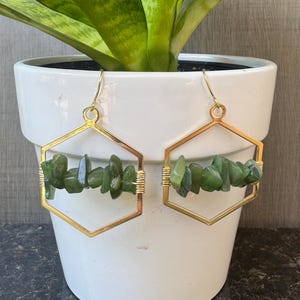 以下が含まれることがあります： ゴールドカラーの六角形イヤリングのペア。各イヤリングは、金のワイヤーで固定された、緑色の不規則な形の石のクラスターが特徴です。イヤリングは、緑色の植物が入った白いポットの前に展示されています。