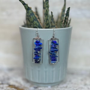 May include: A pair of rectangular silver earrings. Each earring features a vertical stack of blue lapis lazuli chips within a silver frame. The earrings hang from silver hooks. The background includes a light blue pot with a green plant.