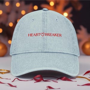 Puede incluir: Gorra de béisbol de mezclilla azul claro con el texto bordado "HEART BREAKER" en rojo. La gorra tiene una visera curva y un diseño de seis paneles. El fondo presenta luces borrosas y elementos decorativos.