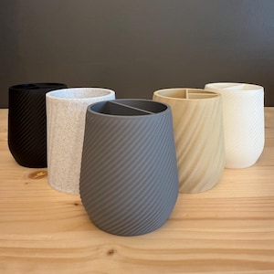 May include: A collection of five cylindrical holders in various colors, including black, speckled white, gray, light wood, and white. Each holder features a diagonal, textured pattern. The holders are arranged on a light wood surface.