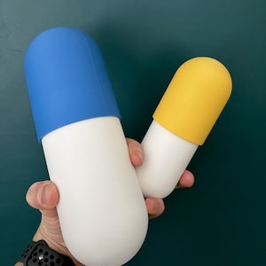 May include: Two pill-shaped containers with smooth, matte surfaces. The larger container features a bright blue cap on a white base, while the smaller container has a yellow cap on a white base. Both items have rounded ends and a cylindrical body.
