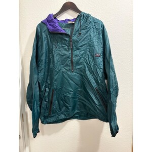 May include: Teal anorak jacket with a purple hood lining and black zipper. The jacket has a half-zip closure, two front pockets, and a small logo on the right chest. The jacket is hanging on a black hanger.