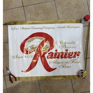 May include: A rectangular white Rainier beer sign with gold and red lettering. The text reads 11 fl.oz. Rainier Brewing Company, Seattle, Washington, Naturally Brewed, Since 1878, and Mountain Fresh Beer. A large red R logo features a mountain graphic.