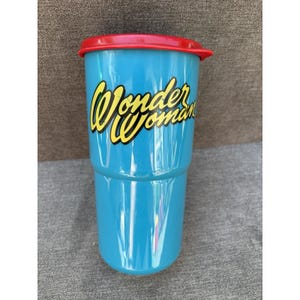 May include: A tall, turquoise tumbler with a red lid. The words "Wonder Woman" are printed in yellow with a black outline on the front of the tumbler. The tumbler is made of a hard plastic material.
