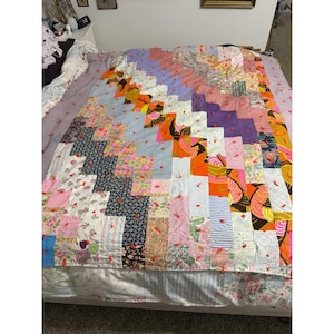May include: A rectangular patchwork quilt featuring a diagonal stair-step pattern of various fabric squares. The quilt displays a mix of floral, paisley, and geometric prints in shades of orange, pink, blue, yellow, and white, accented with small red ties.